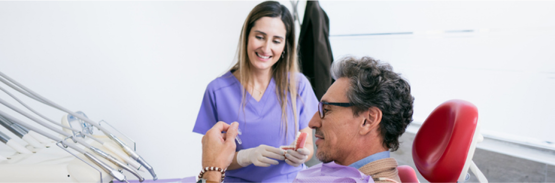 Dentista com uniforme roxo explicando um tratamento para paciente idoso sentado em cadeira odontológica em consultório moderno.